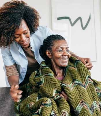 Caregiver wrapping a warm blanket around an older woman with dementia at home.