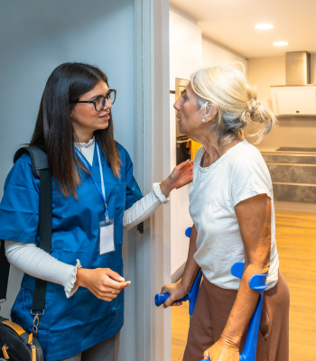 Caregiver guiding an older woman with crutches through a home doorway during a personal care visit.