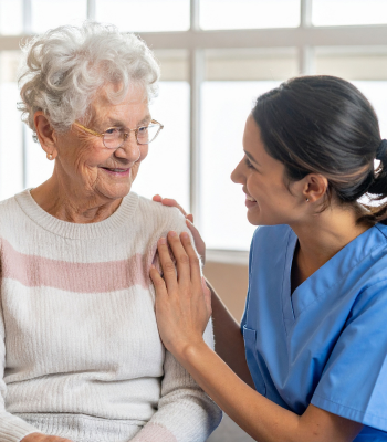 Caregiver in blue scrubs gently resting a hand on an older woman's shoulder during a home visit.
