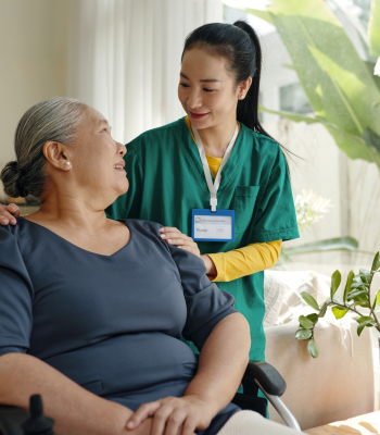 Caregiver in green scrubs reassuring an older woman seated in a wheelchair in her sunlit living room.